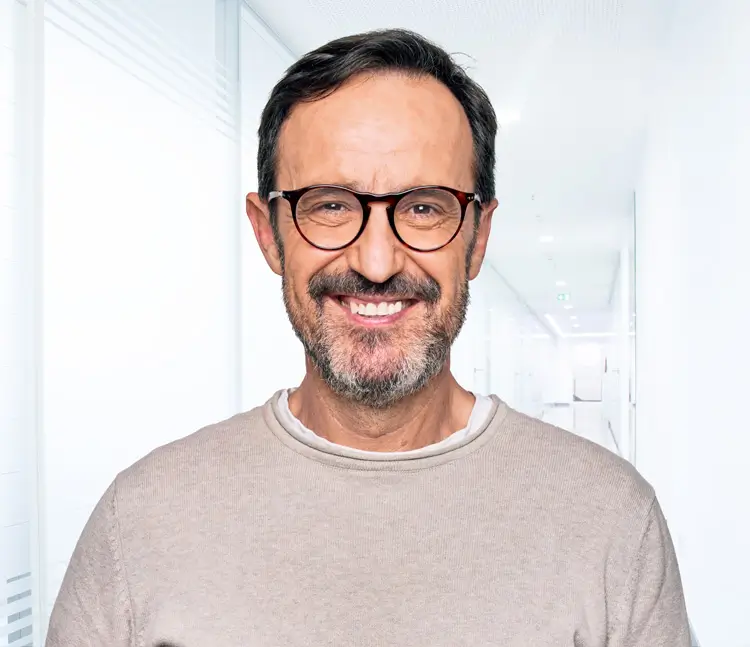 Portr&auml;t eines l&auml;chelnden Mannes mit Brille, Bart und hellem Pullover vor grauem Hintergrund