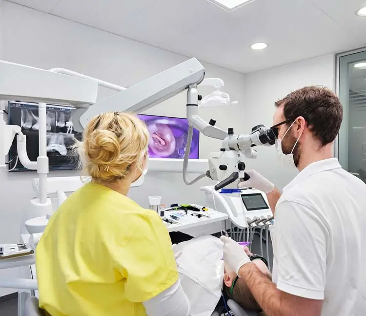 Zahnarzt mit Assistenz arbeiten an einem Patienten. Einer benutzt ein Mikroskop, w&auml;hrend die andere danebensteht. Im Hintergrund sind R&ouml;ntgenbilder und ein Monitor zu sehen.