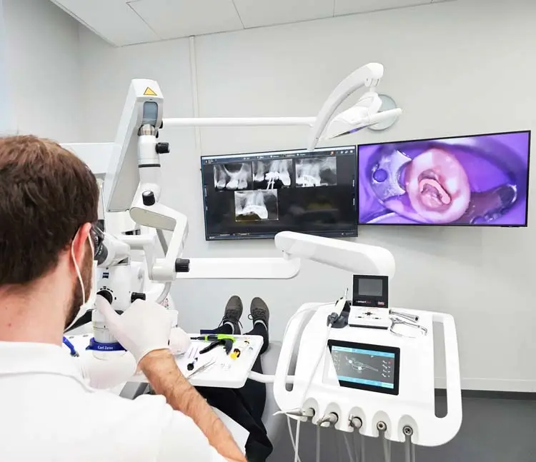 Ein Zahnarzt arbeitet mit einem Mikroskop an einem Patienten. Im Hintergrund sind Monitore mit R&ouml;ntgenaufnahmen und einer Nahaufnahme eines Zahns zu sehen