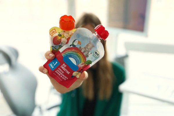 Person h&auml;lt zwei bunte Quetschbeutel mit Bio-Smoothies in die Kamera, im Hintergrund unscharf eine Zahnarztpraxis